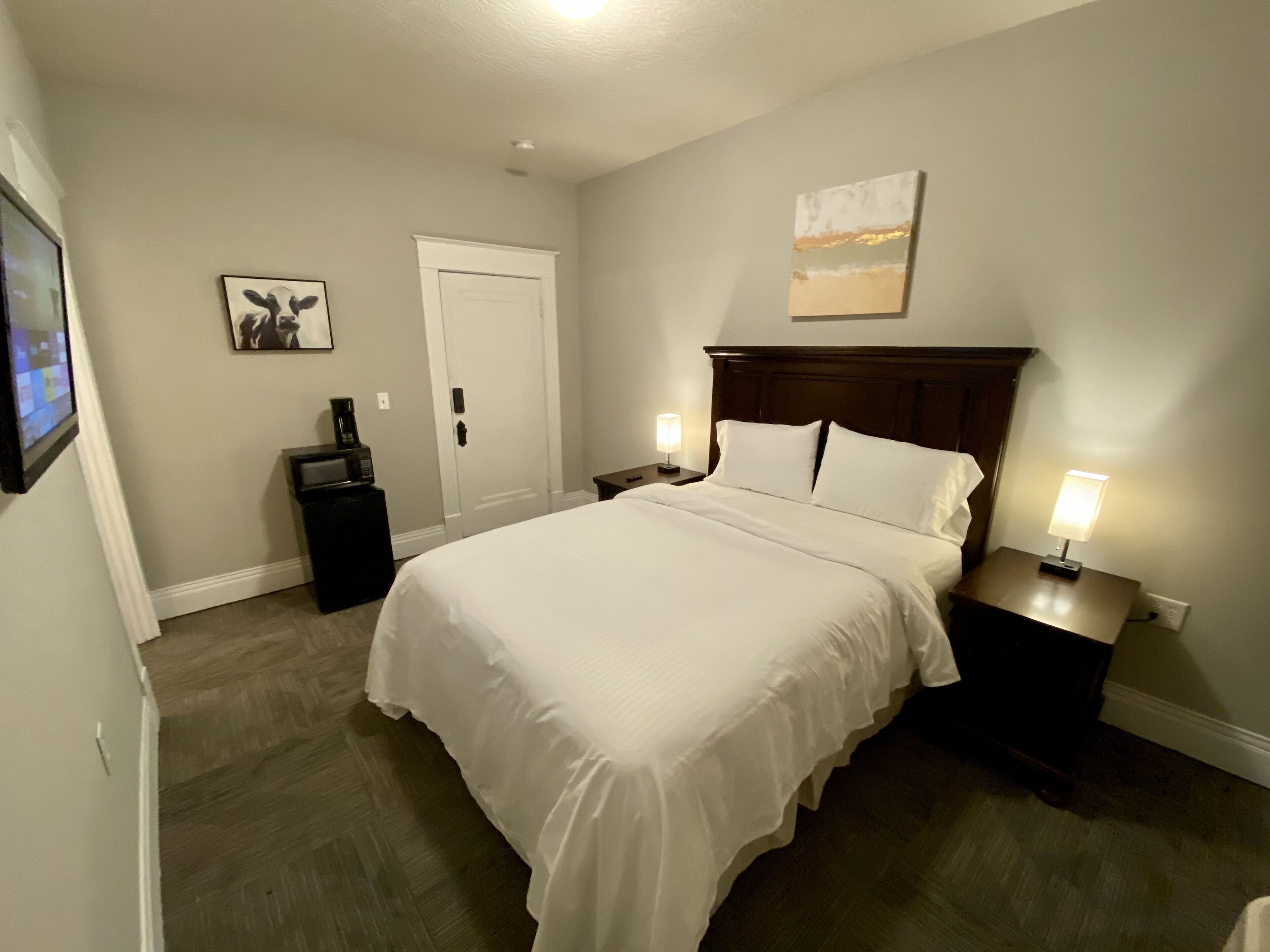 Une chambre d’hôtel avec un grand lit, deux tables de chevet avec des lampes, une porte, un tableau encadré au mur et une petite table sur laquelle se trouve une machine à café.