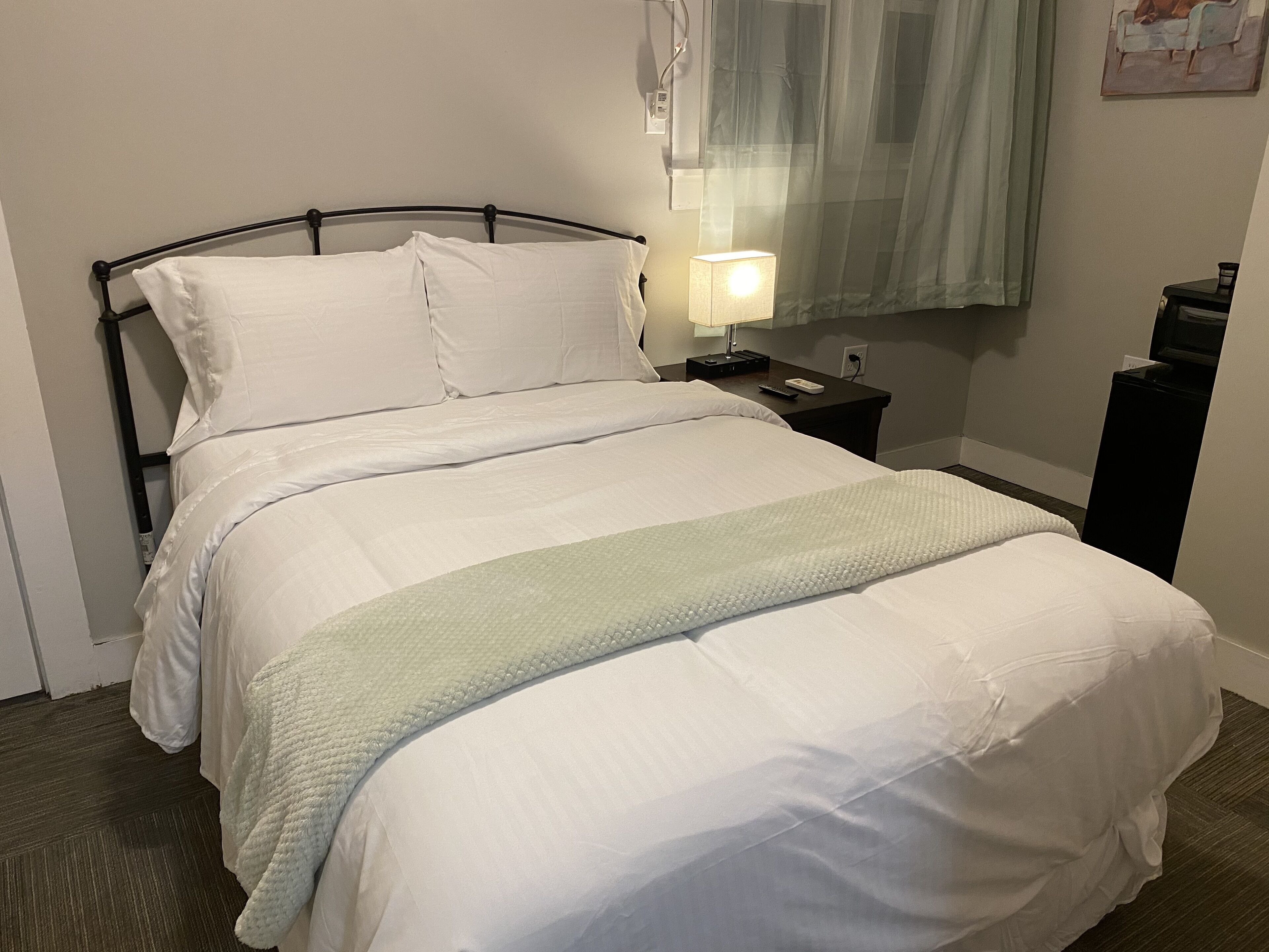 A neatly made bed with white linens and a light green throw blanket. There is a bedside table with a lamp and a phone. The room has a simple, clean design with a window covered by a sheer curtain.