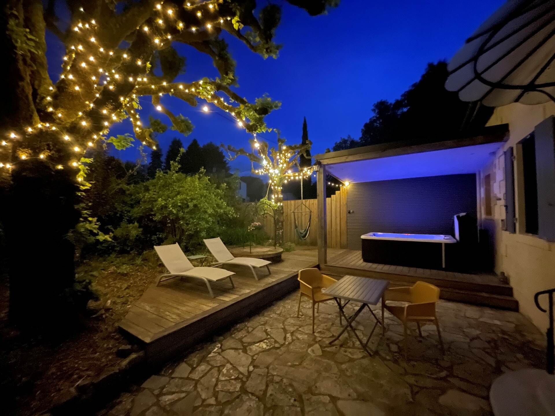 An outdoor patio with stone flooring, two lounge chairs, a hot tub, and string lights.