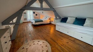 Une chambre mansardée chaleureuse avec un plafond en pente, un plancher en bois, un lit avec une literie blanche, un canapé blanc avec des coussins bleus et une petite commode blanche.
