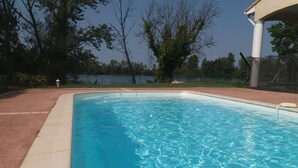 Piscina clara com borda azulejada, cercada por área ajardinada com árvores e cerca.