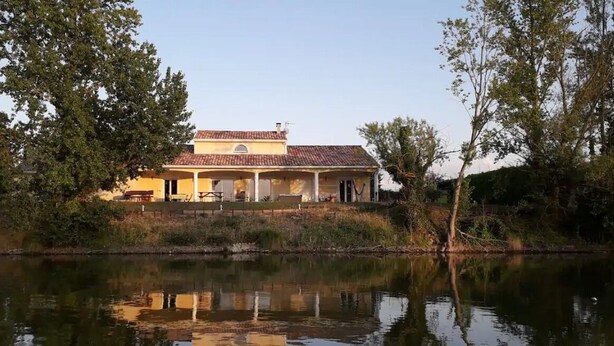Uma casa de dois andares com telhado de telhas vermelhas, varanda coberta e um lago em primeiro plano.