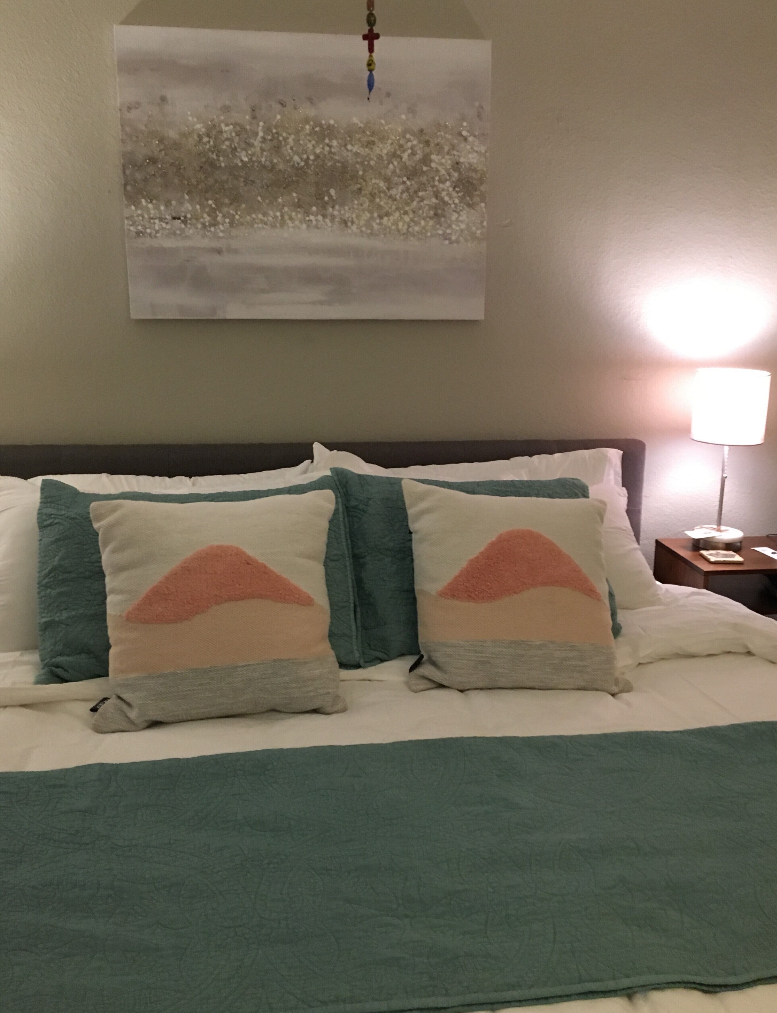 Un letto con una coperta verde acqua, due cuscini con motivo montano e un quadro appeso al muro raffigurante un paesaggio innevato.