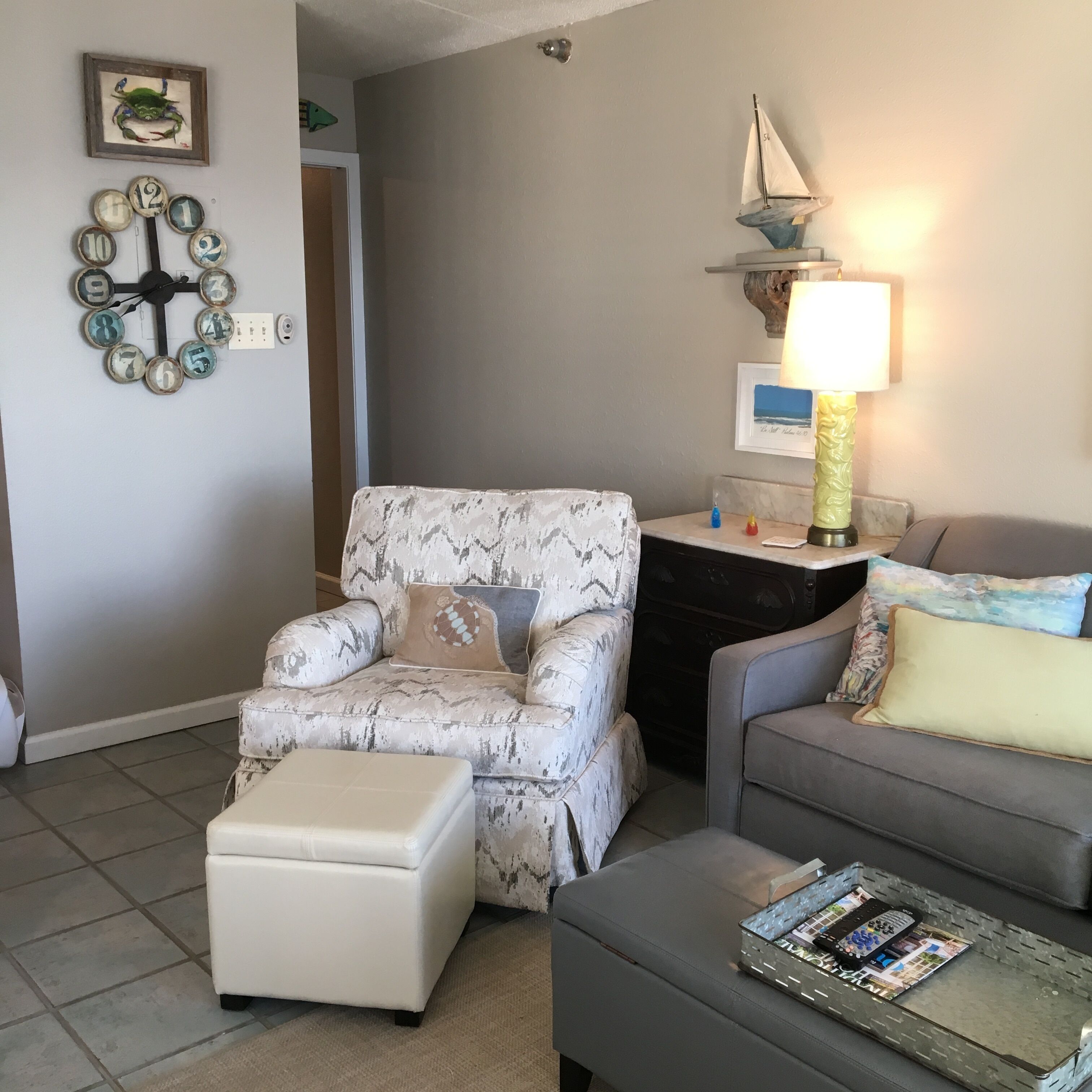 A living room with a patterned armchair, a grey sofa, and a white ottoman. There is a wall clock with numbers, a framed picture of a crab, and a small sailboat decoration.