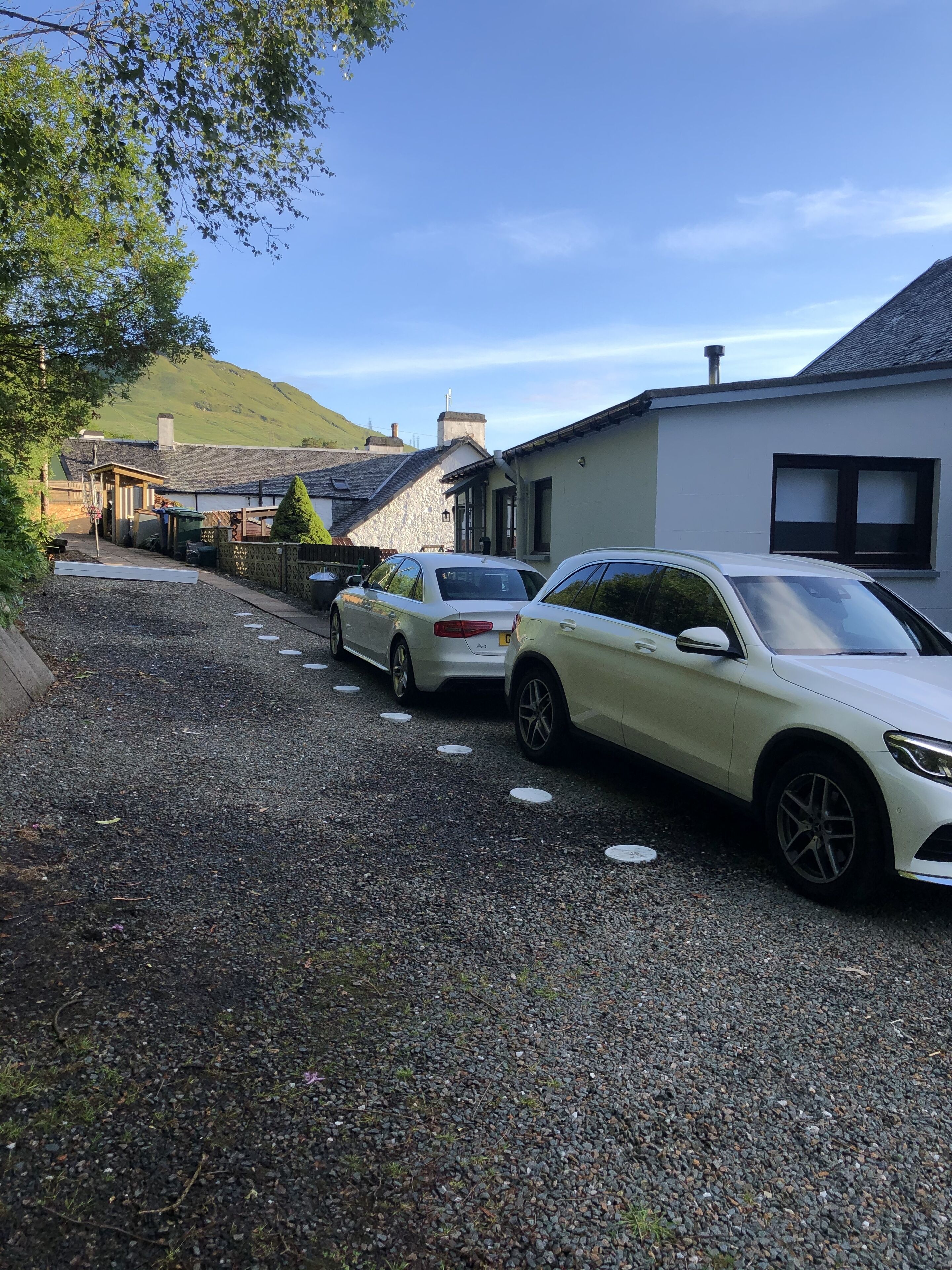 A gravel driveway with two parked cars, a stone building, and a hill in the background.