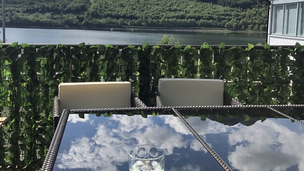 Een glazen tafel met een helder glas water, omringd door rieten stoelen, met uitzicht op een meer en bergen.