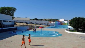 A swimming pool area with a water fountain, a beach in the background, and a modern building with a colorful facade.