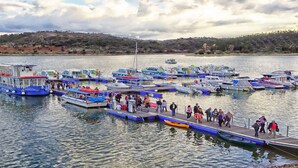 A marina with multiple boats docked, a pier with people walking, and a hilly landscape in the background.
