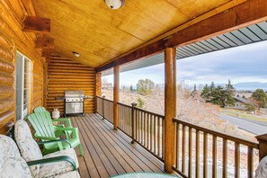 Una baita di legno con un portico, sedie Adirondack verdi, un barbecue e vista su una strada e alberi.