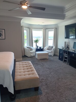A hotel room with a bed, a desk, a chair, a small table, a ceiling fan, and a large window with curtains.