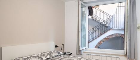 Un dormitorio con una cama grande, una ventana que da a una escalera y ladrillo, y un radiador debajo de la ventana.