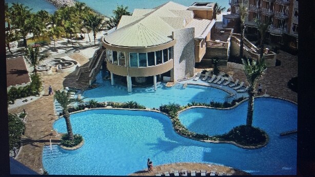 A resort with a central circular pool, a palm tree-lined walkway, and a multi-story building with balconies. - 5 Star Aruba Resort Christmas week! (Noord)