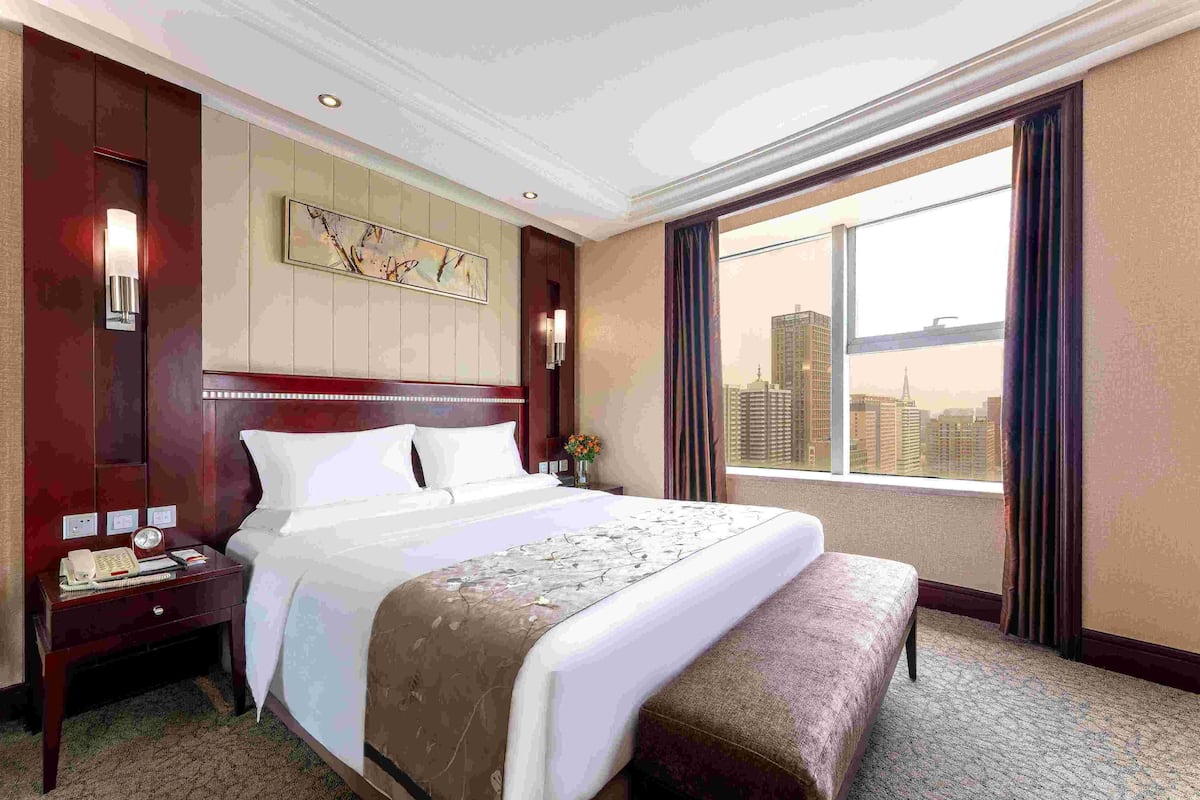 A hotel room with a large bed, a nightstand with a phone and notepad, a window with a city view, and a decorative wall piece.