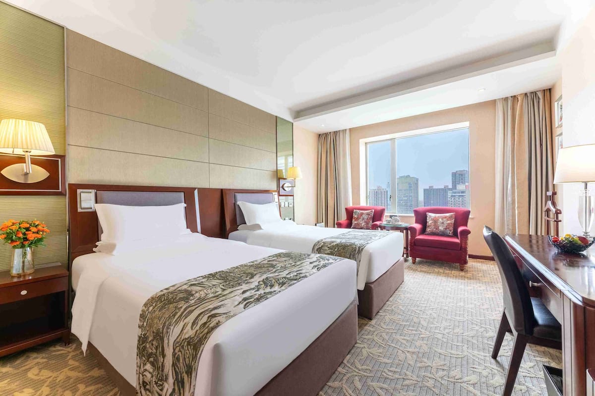 A hotel room with two beds, a desk, a chair, and a large window offering a city view.