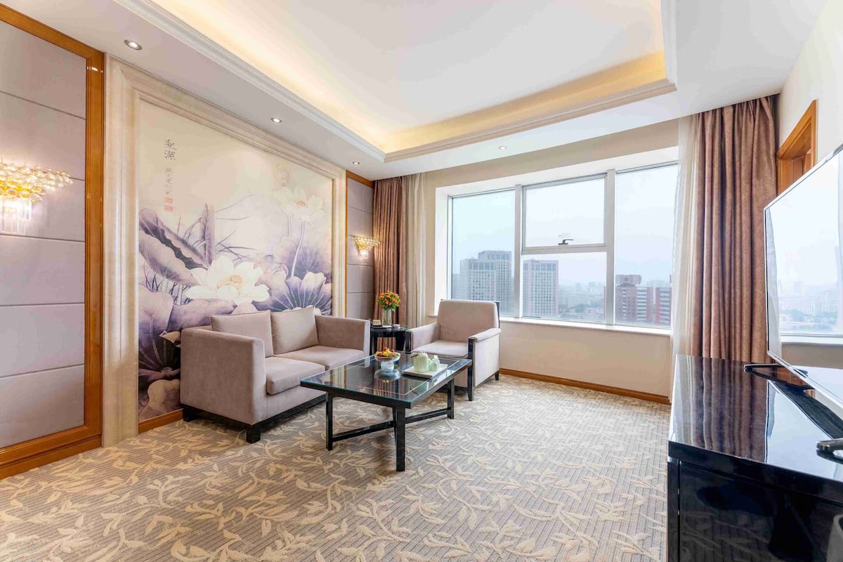 A modern hotel room with a large window, a sofa, a coffee table, and a TV.