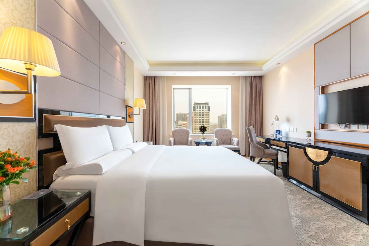 A hotel room with a large bed, a desk with a chair, a TV, and a view of the city through the window.