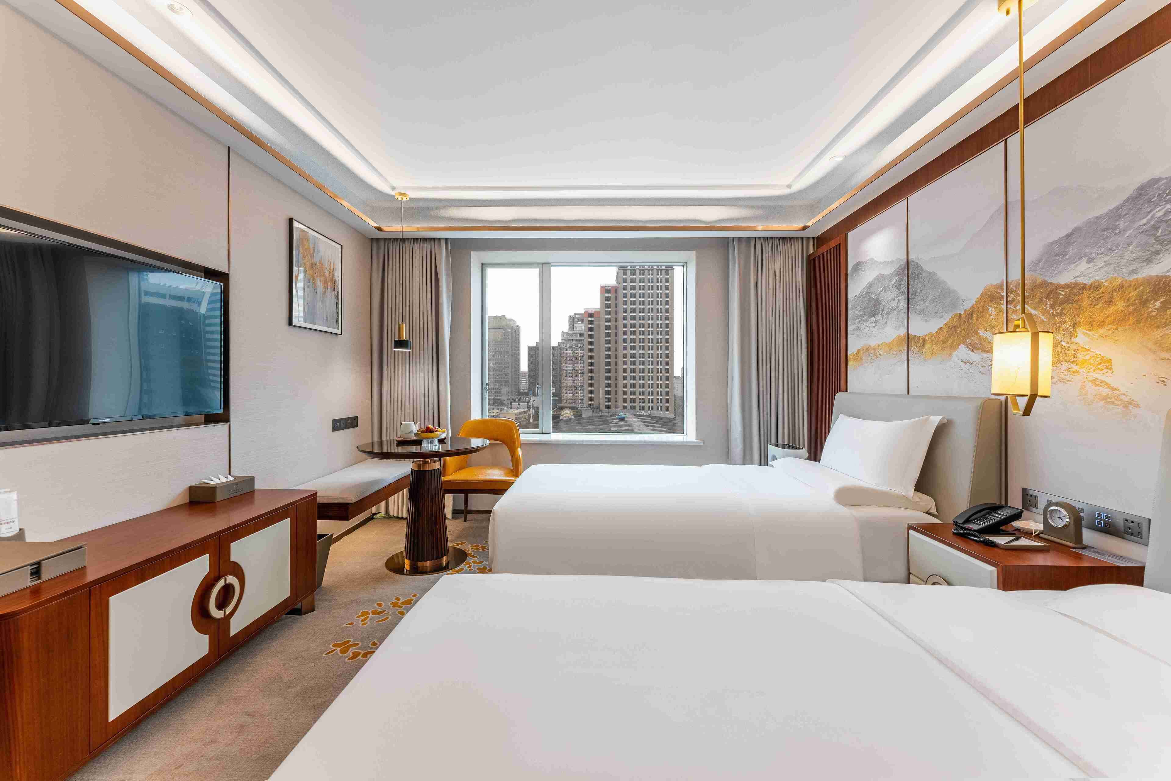 A hotel room with two beds, a desk, a chair, a TV, a window with a city view, and a painting on the wall.