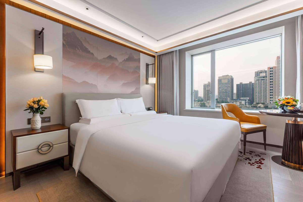 A modern hotel room with a large bed, a desk with a chair, a city view, and a decorative painting on the wall.