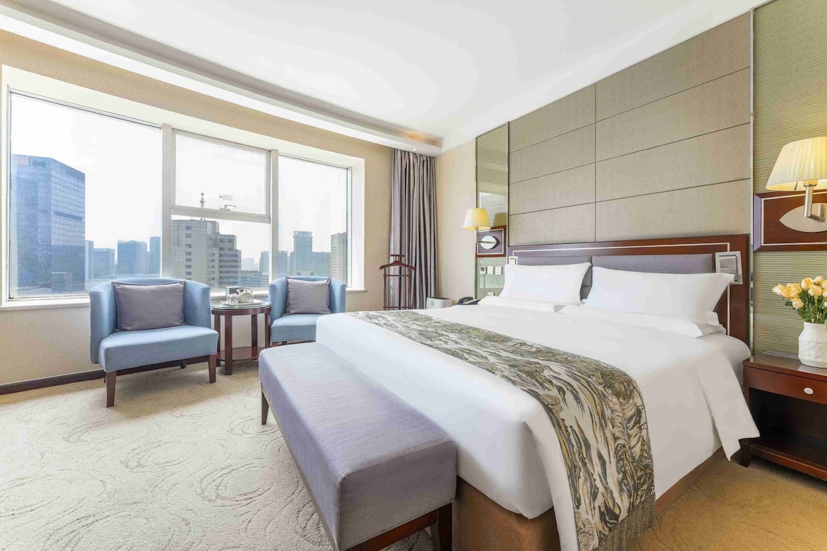 A hotel room with a large bed, a seating area with a chair and a bench, a small table, and a view of the cityscape through large windows.