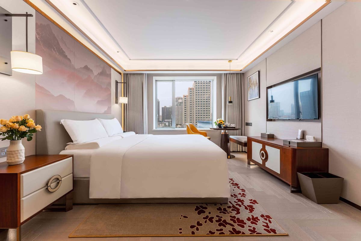 A modern hotel room with a large bed, a desk, a chair, a TV, and a view of the cityscape.