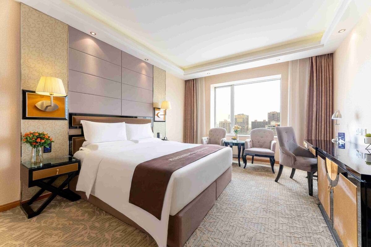 A hotel room with a large bed, a desk, two chairs, a city view, and a decorative vase with flowers.