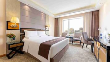 A hotel room with a large bed, a desk, two chairs, a city view, and a decorative vase with flowers.