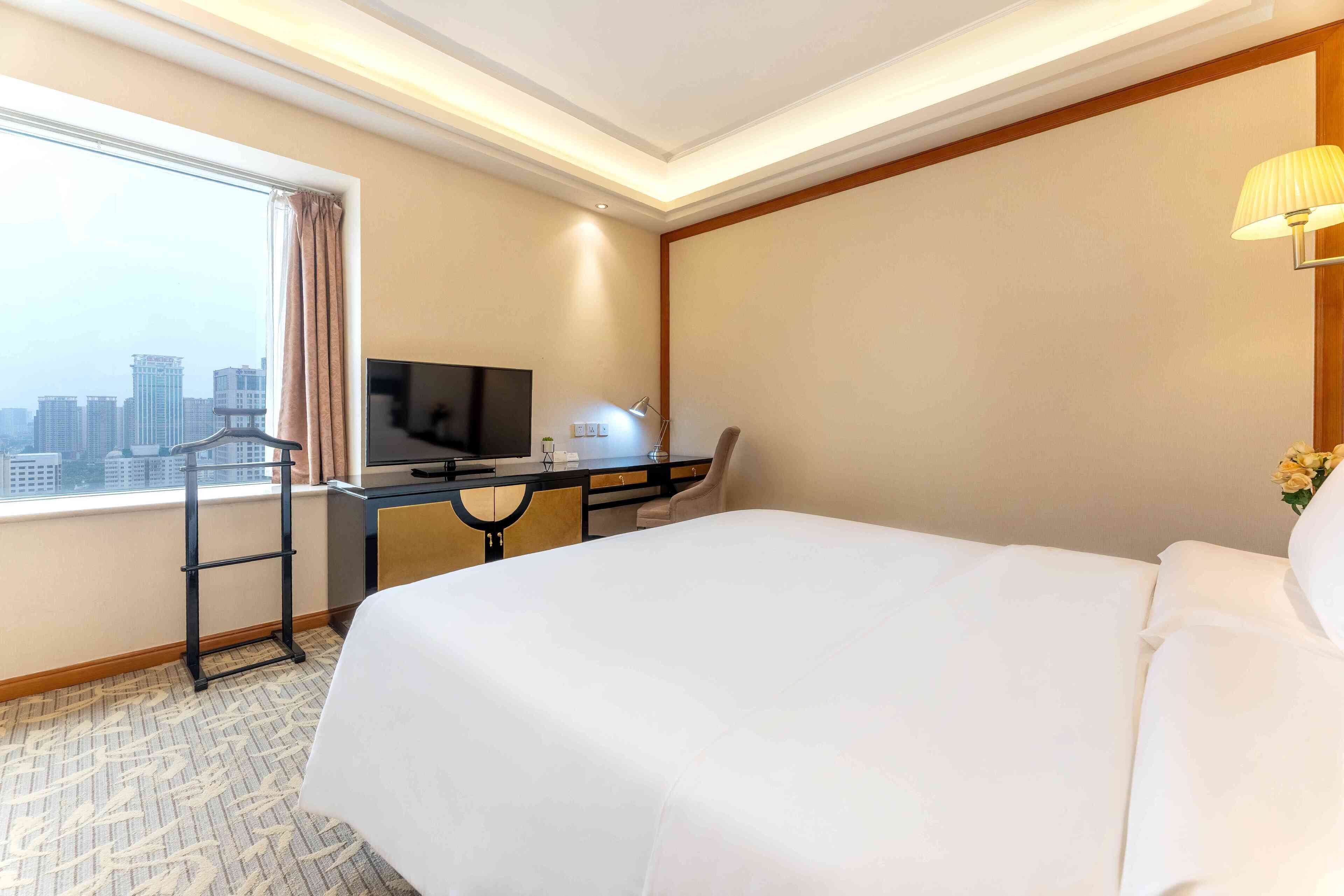 A hotel room with a large bed, a desk with a computer and lamp, a TV, and a window with a city view.
