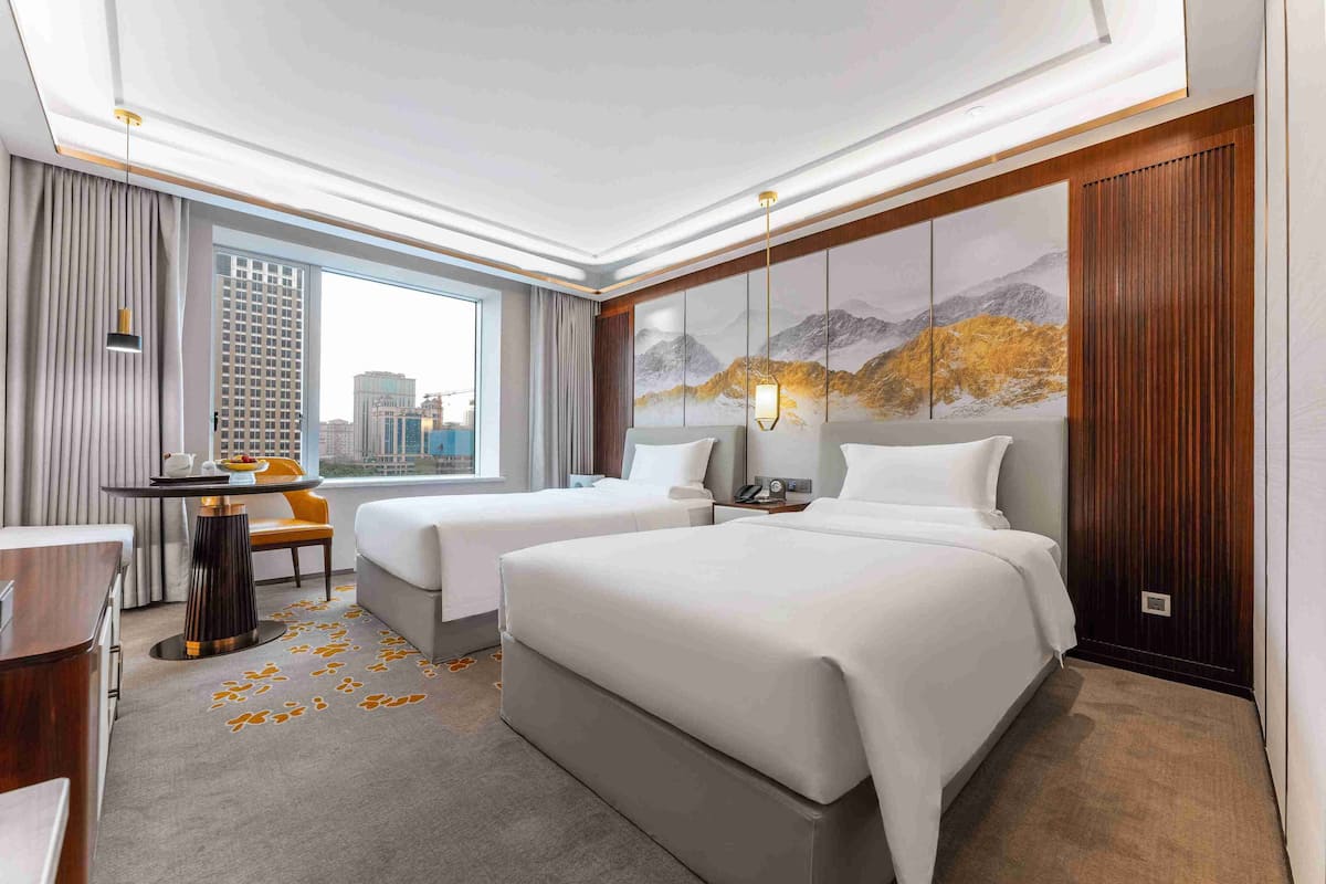 A hotel room with two beds, a desk, a chair, and a large window offering a city view.