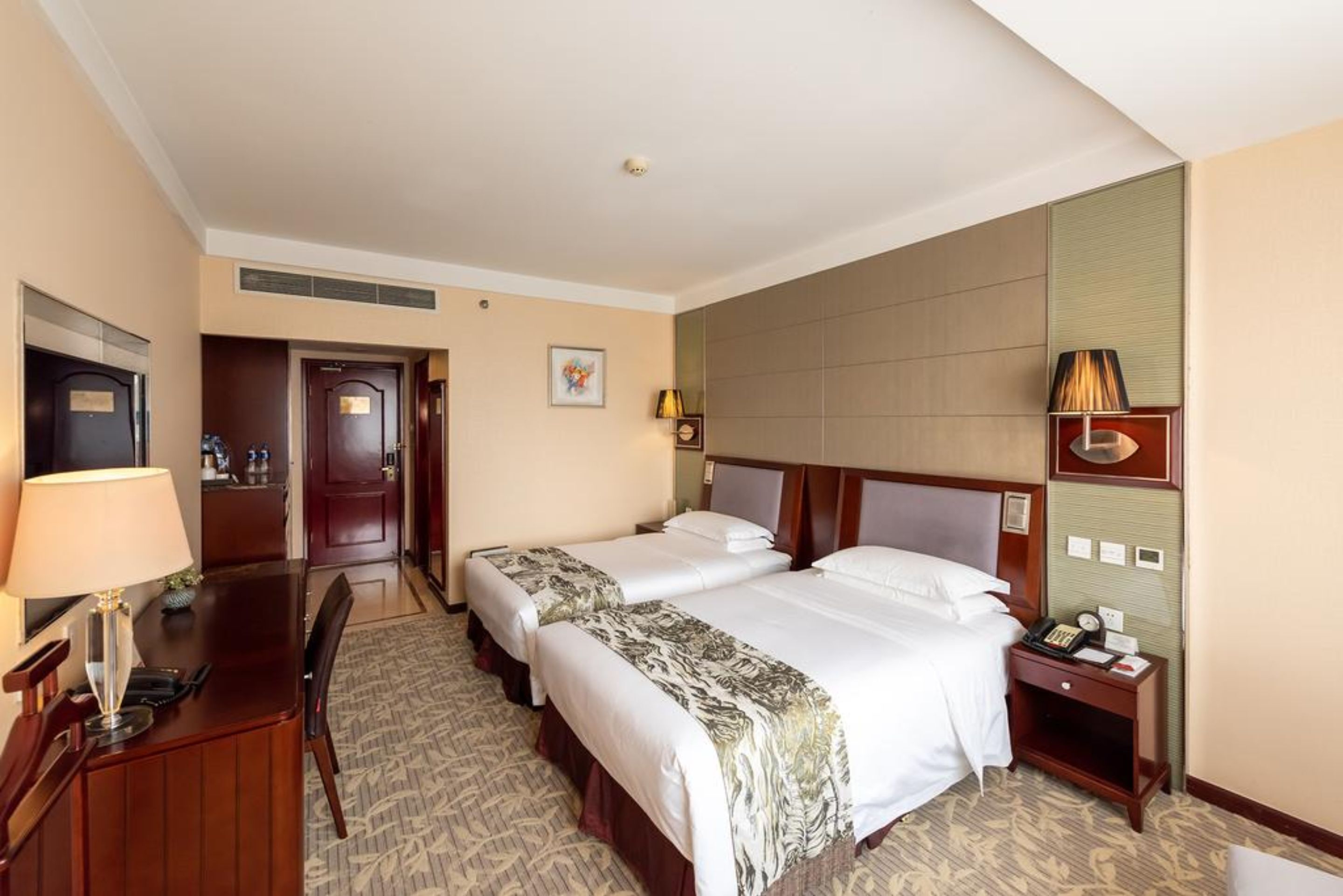 A hotel room with two beds, a desk, a chair, a lamp, a nightstand, and a wall-mounted telephone.