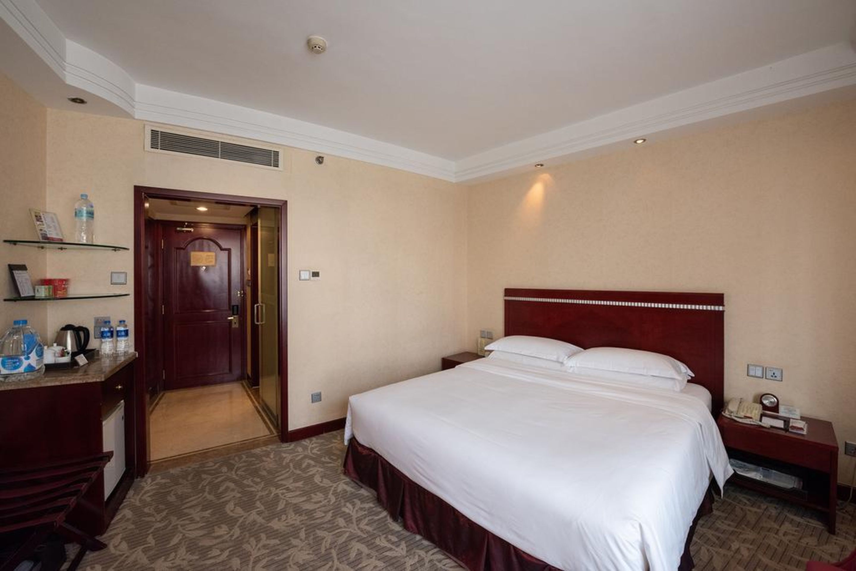 A hotel room with a large bed, a desk with a chair, a nightstand, and a shelf with toiletries.