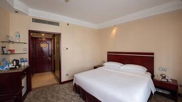A hotel room with a large bed, a desk with a chair, a nightstand, and a shelf with toiletries.