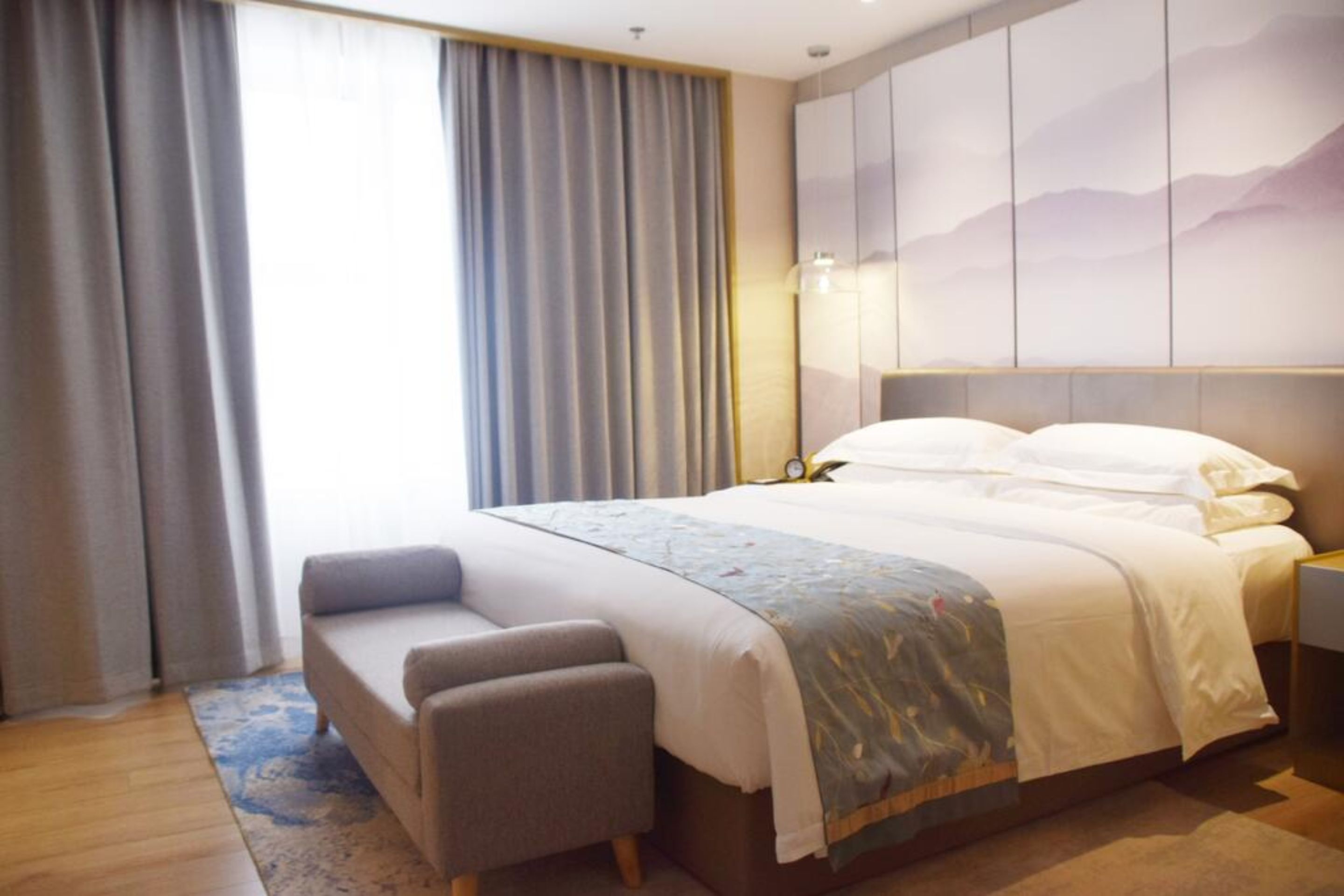 A hotel room with a large bed, a bedside table, a chair, and a view of the outdoors through the window.