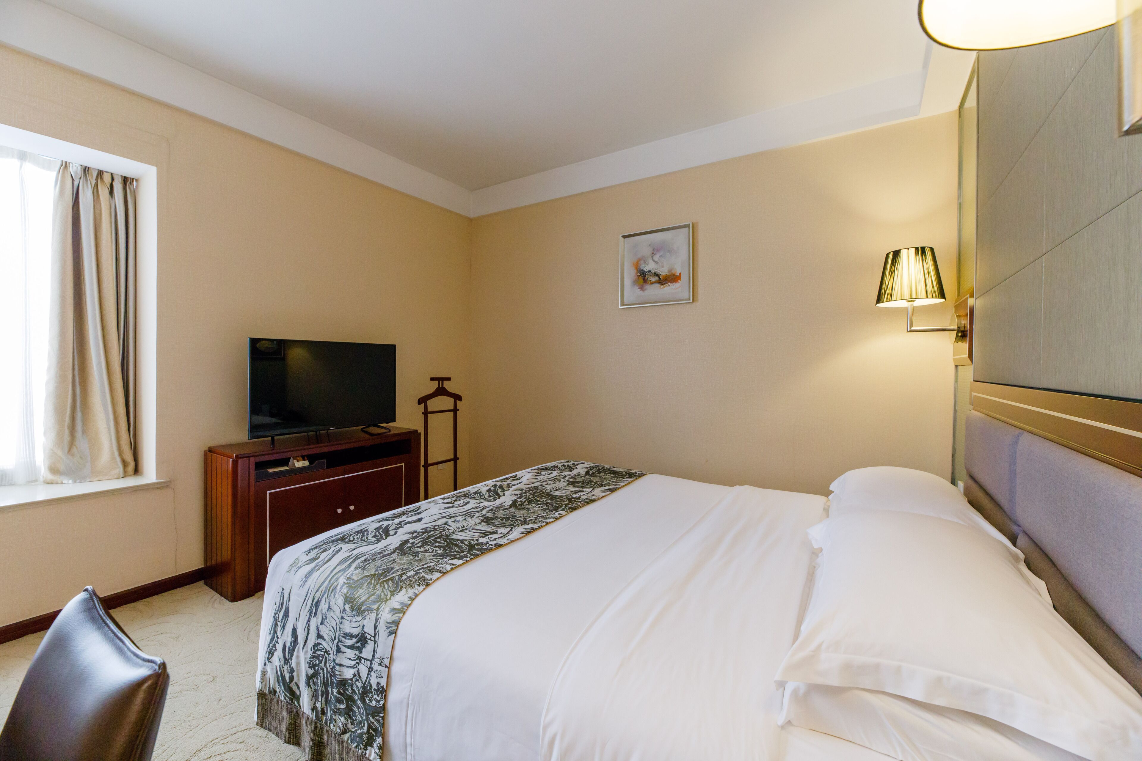 A hotel room with a bed, a television on a wooden cabinet, a lamp, and a window with curtains.