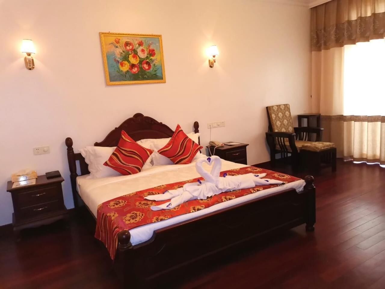 Ein ordentlich bezogenes Bett mit roter und goldener Bettdecke, zwei Wandlampen, ein Blumenbild und ein Sessel in einem Hotelzimmer.