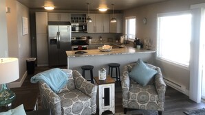 A modern kitchen with stainless steel appliances, a central island, and pendant lighting. There are two patterned armchairs and a small table with a lamp. - Edgewater #8 Scout (Polson)