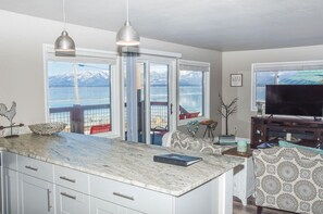 Cocina con una isla grande, dos lámparas colgantes, vista a un lago con montañas y un comedor con tapicería estampada.