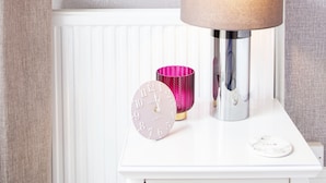 A white bedside table with a lamp, a pink clock, and a decorative mirror on the wall above. - The Rosewood Torquay (Torquay)
