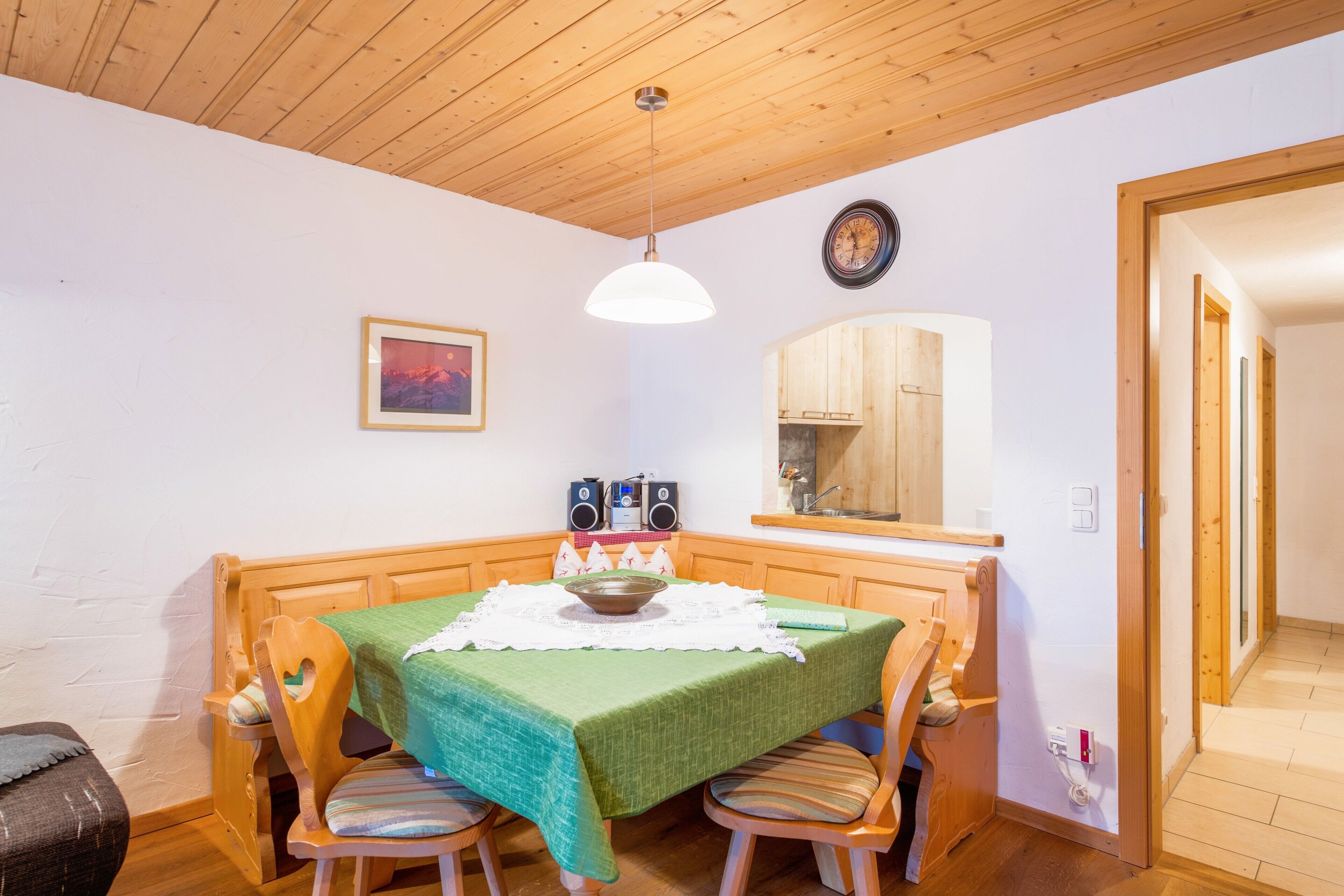 Ein Essbereich mit Holztisch, Stühlen, einer grünen Tischdecke, einem weißen Spitzenläufer, einer hängenden Pendelleuchte, einer Uhr und einem gerahmten Bild an der Wand.