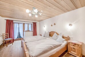 Quarto com cama de madeira, uma cadeira, mesa de cabeceira e janela com cortinas.