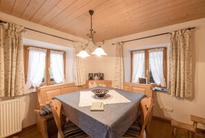 Een gezellige kamer met een houten plafond, een eettafel met een schaal en een boek, en twee ramen met gordijnen.
