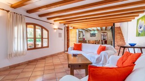Un vaste espace de vie doté d’un plafond en bois, de canapés blancs agrémentés de coussins orange, d’une table en bois et offrant une vue sur la cuisine et l’escalier.