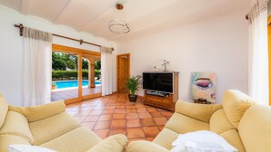 A living room with a TV, sofa, and a view of a swimming pool. - “Ca Na Rosa” close to the Beach with Wi-Fi, A/C, Terrace, Garden & Pool (ALCUDIA)