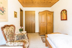 A bedroom with a wooden wardrobe, a floral armchair, a bed, and a door leading to another room. - Cosy Single Apartment in Spa Town in the Southern Black Forest (Todtmoos)