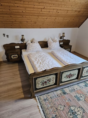 Un dormitorio con techo de madera, una cama con cubrecama floral, dos mesitas de noche con lámparas y una alfombra estampada.