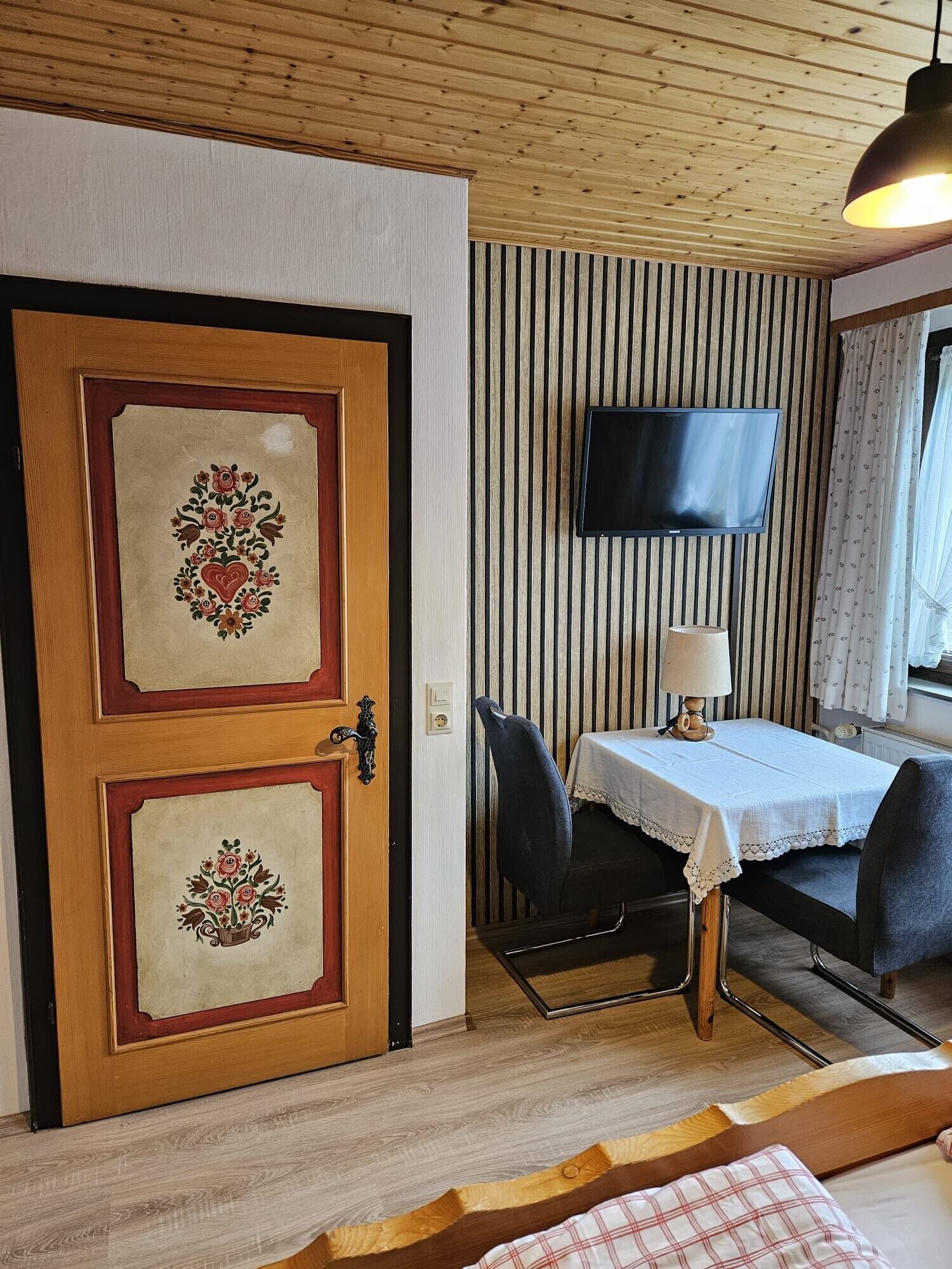 Una habitación con puerta de madera, televisor montado en una pared a rayas, una mesa con sillas y una cama con manta escocesa.