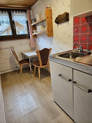 Une cuisine avec une table en bois, deux chaises, un évier et une fenêtre ornée de rideaux en dentelle.