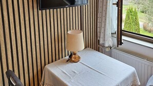 A room with a TV mounted on a wooden wall, a table with a white tablecloth, and a window with a view of greenery. - Apartment "Am Wehrastrand 5" in a Spa Town in the Southern Black Forest with Mountain View (Todtmoos)