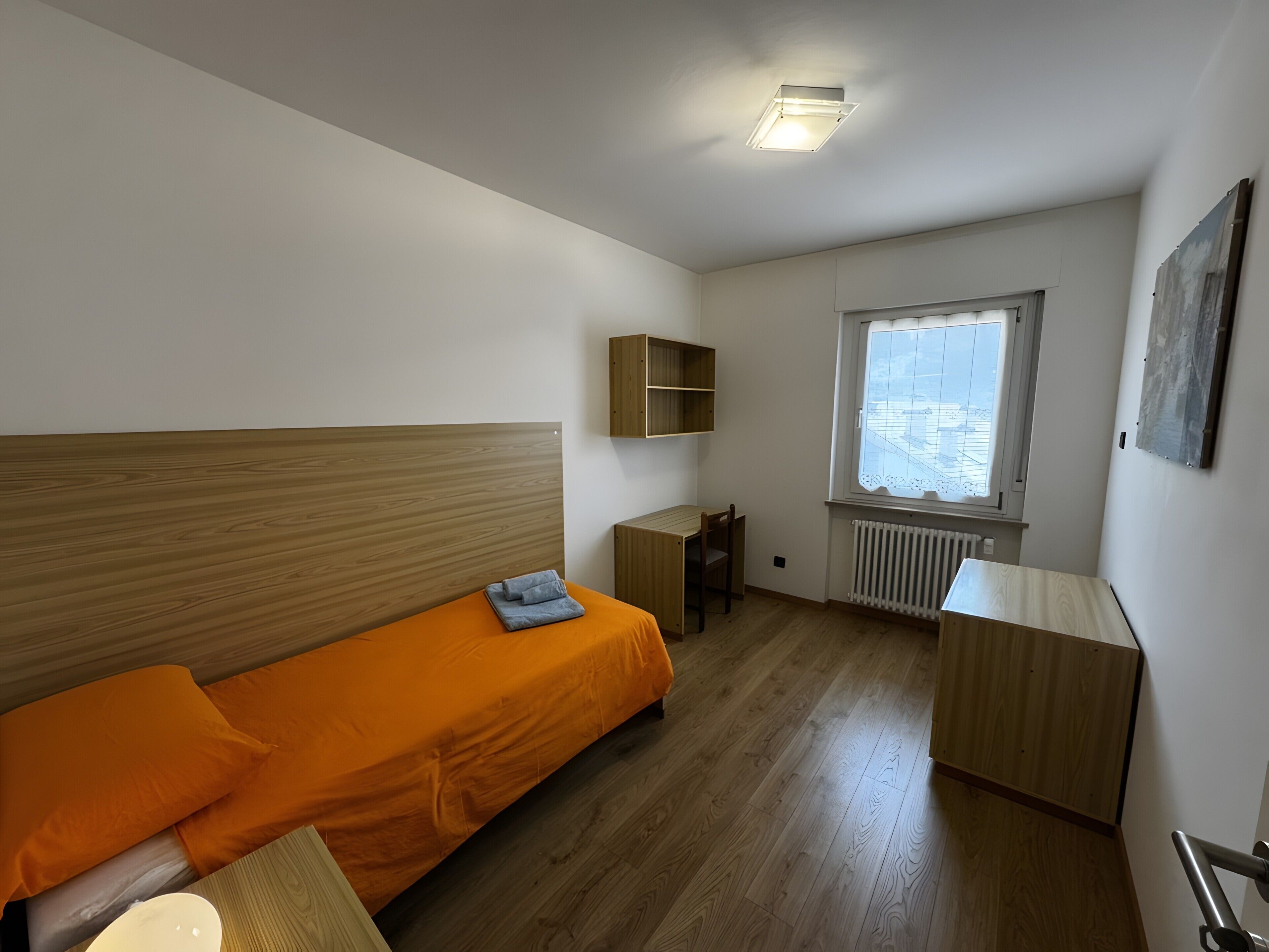 Quarto individual com cama de solteiro, cabeceira de madeira, colcha laranja, pequena escrivaninha de madeira, radiador e janela com vista.