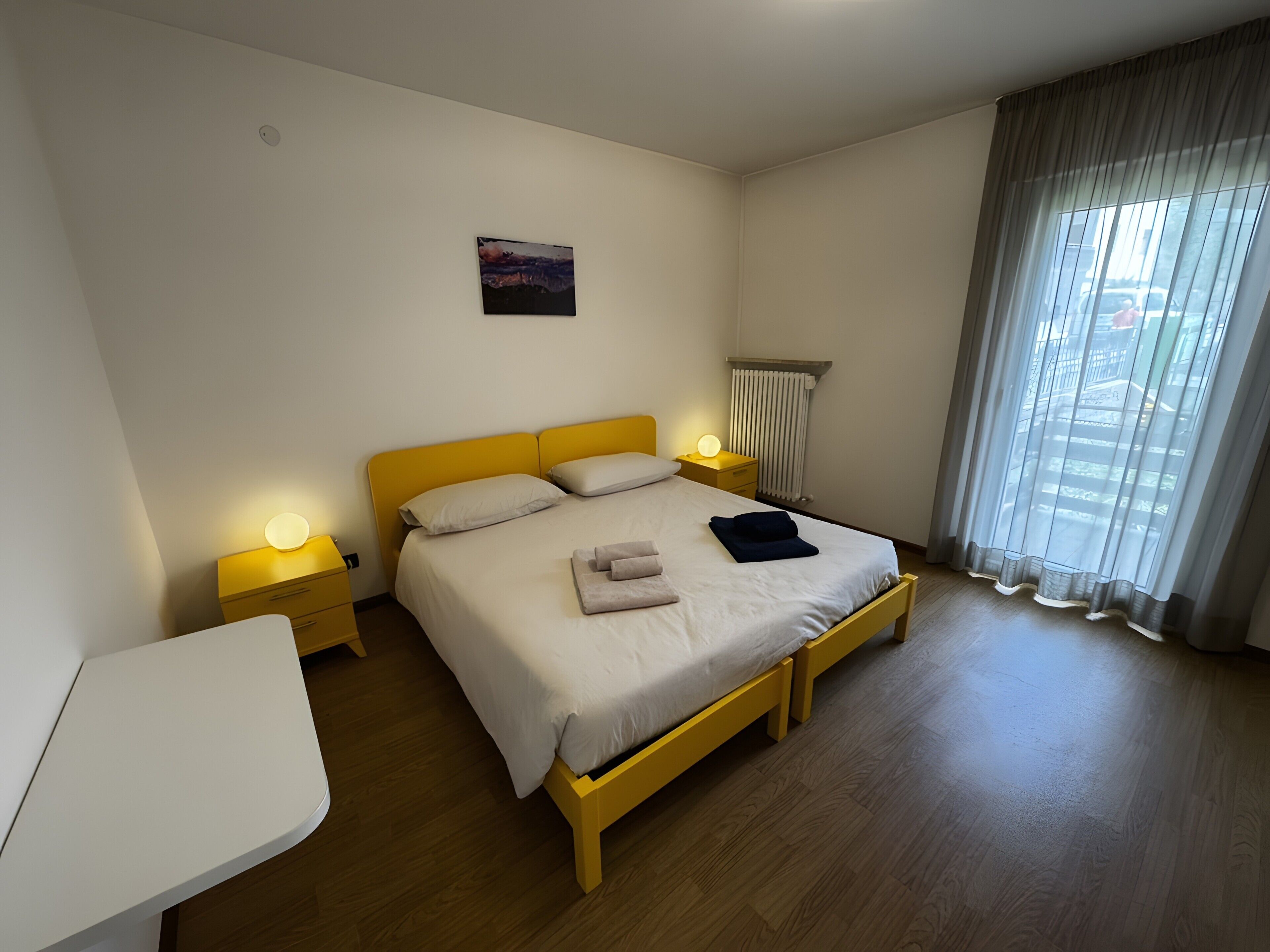 Une chambre d’hôtel comprenant un lit, des tables de chevet, une commode, une fenêtre avec des rideaux et un tableau accroché au mur.