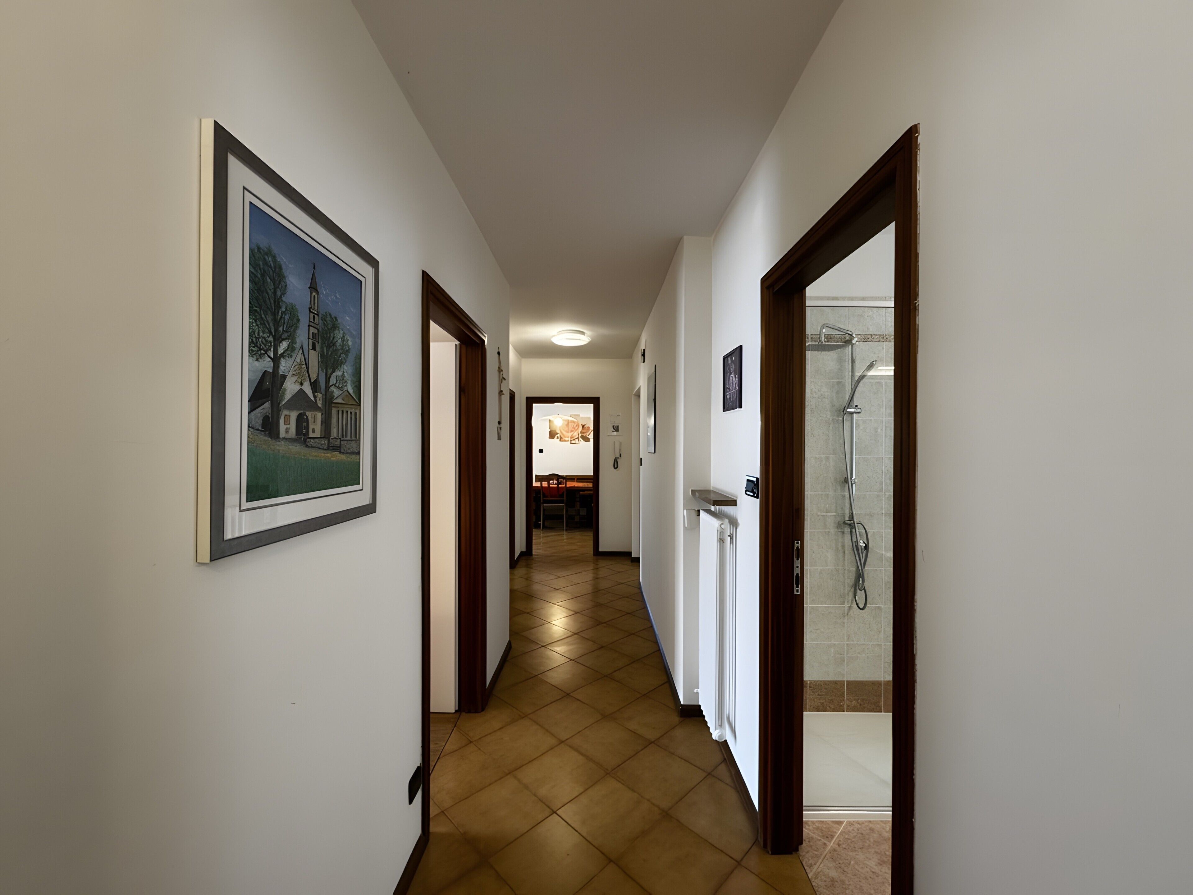 A hallway with a framed picture on the left, a shower area on the right, and a dining area in the background.
